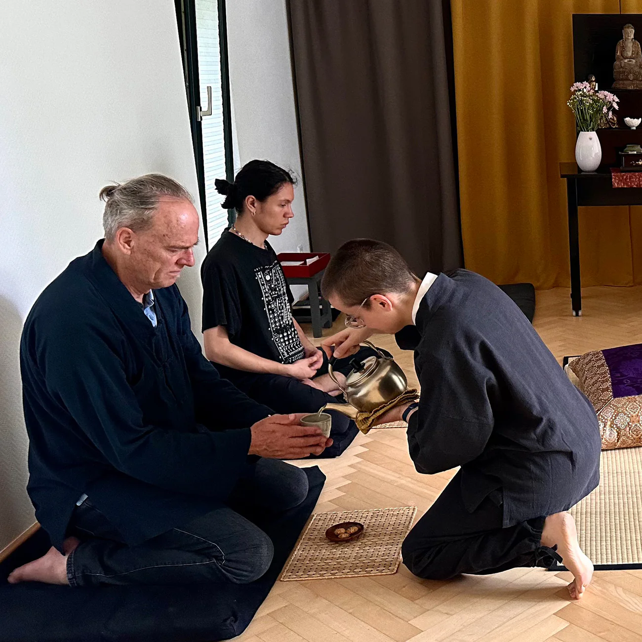 Traditionelles japanisches Teeritual in einem Zendo: Eine kniende Person gießt Tee aus einem metallenen Wasserkessel (Tetsubin) in die Schale eines sitzenden Mannes. Im Hintergrund meditiert eine weitere Person im Lotussitz vor einem Altar mit Buddha-Statue.